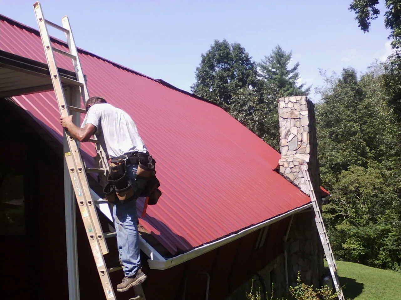 Roofer on a ladder beside a red metal roof and stone chimney — Soffit & Fascia Repair by Roofing Brothers in Melvern